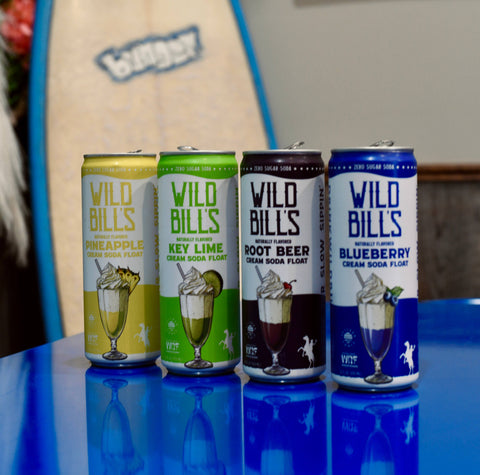 Four cans of Wild Bills cream soda float flavors on a reflective surface with a blurred background.
