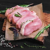 Raw pork chops on a wooden board with herbs and spices on a dark surface