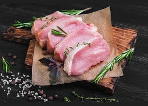 Raw pork chops on a wooden board with herbs and spices on a dark surface