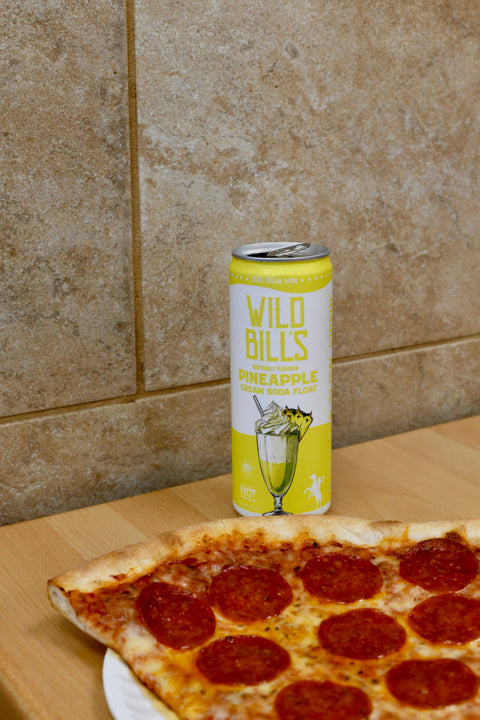 Can of Wild Bills Pineapple Cream Soda on a wooden surface with a pizza in the foreground.