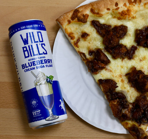 Can of Wild Bills Blueberry Cream Soda Float next to a slice of pizza on a wooden table.